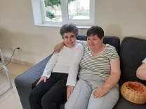 Márta umarmt Erika. Beiden sitzen auf einer Couch im Haus des Lächelns.